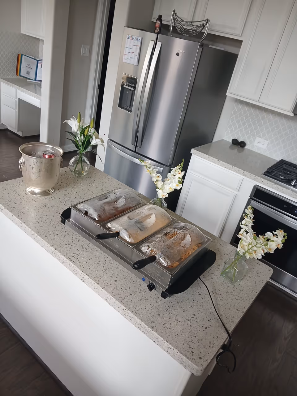 Modern kitchen with island, stainless fridge, white cabinets, and decorative flowers on the counter.