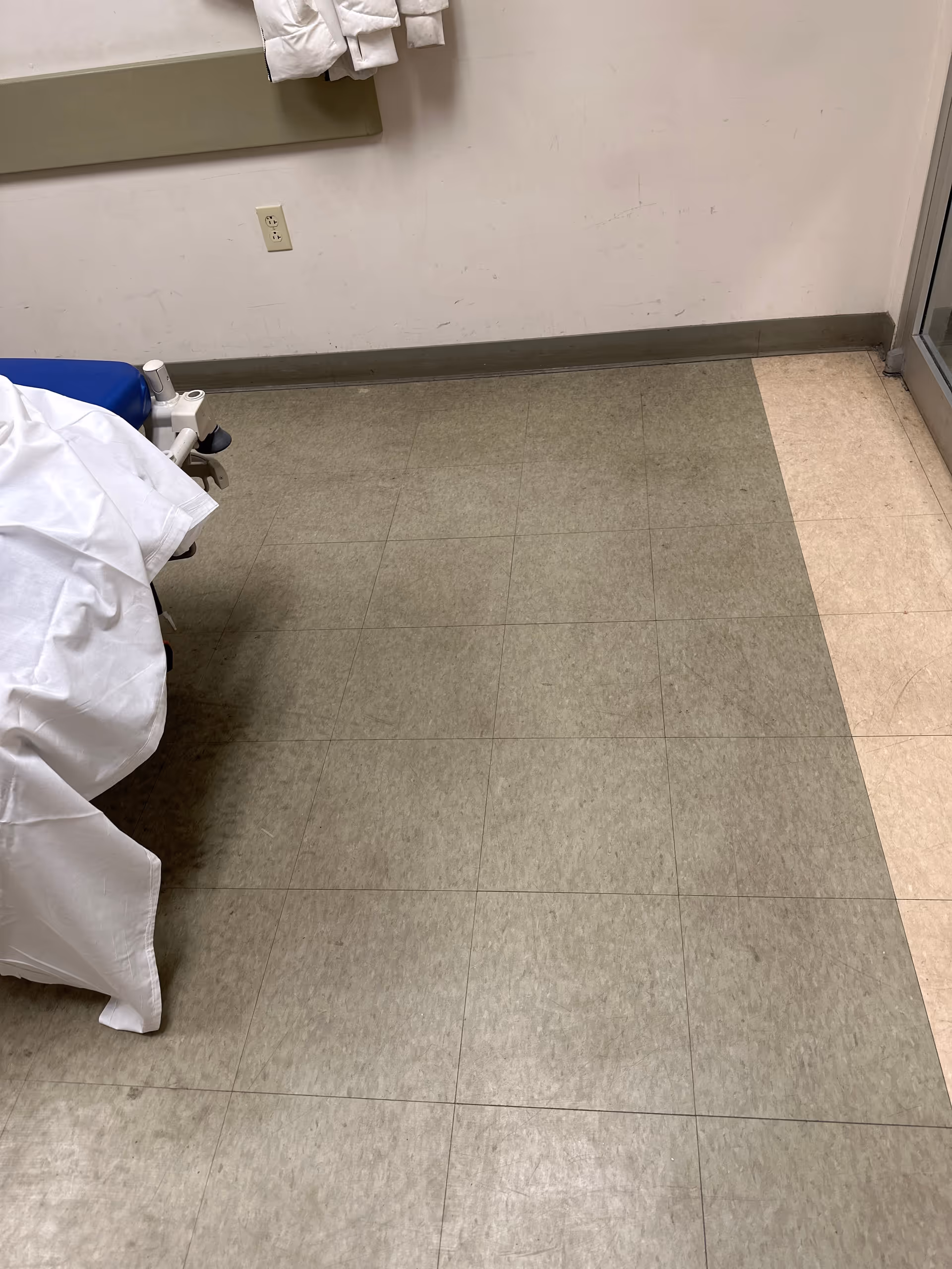 Hospital room corner with a bed, coats hanging, and tiled floor.
