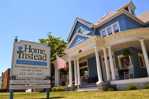 Blue colonial-style senior care home with a front porch and Home Instead sign.