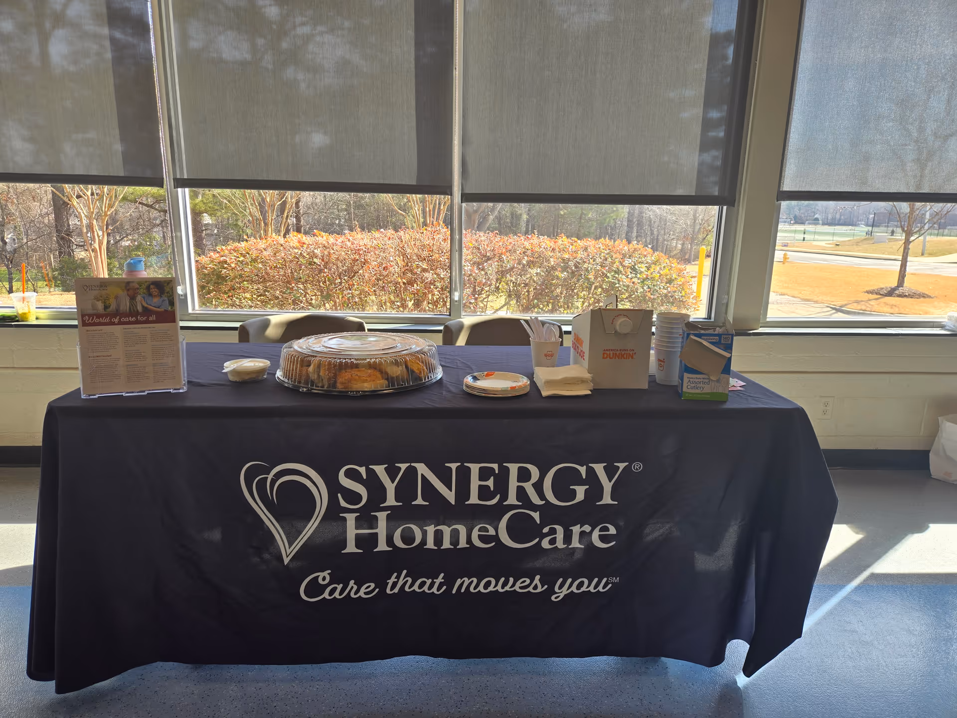 Indoor table with SYNERGY HomeCare tablecloth, cake, cups, and napkins by windows.