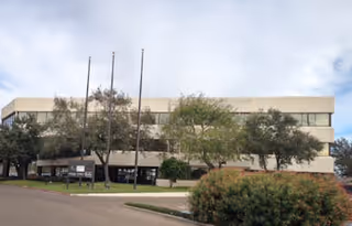 Exterior view of a two-story beige building with trees, shrubs, and flagpoles in front.