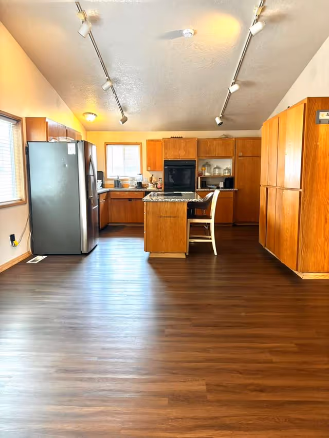 Spacious kitchen with wooden cabinets, island, stainless steel refrigerator, and dark wood flooring.