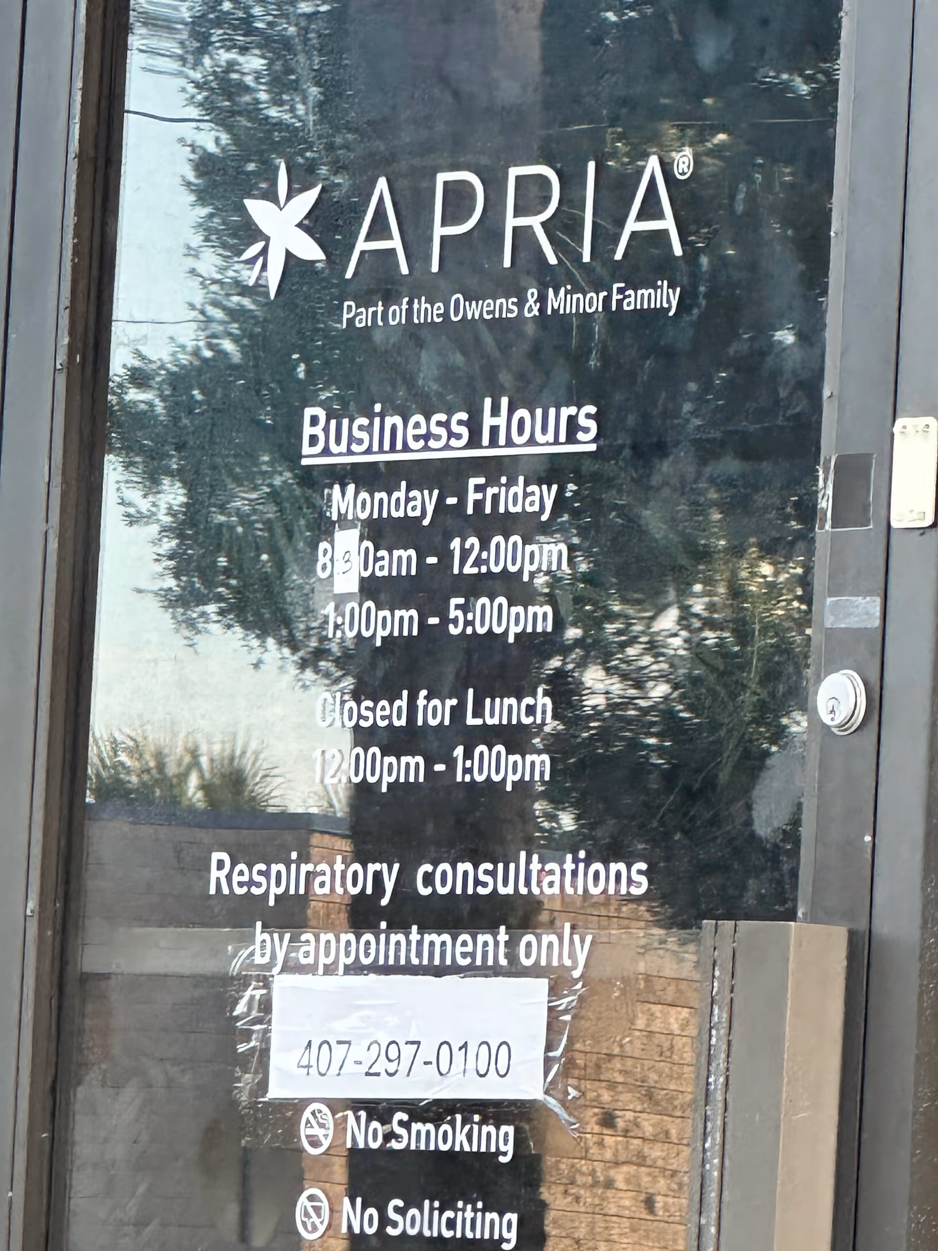 Glass door shows Apria logo and business hours, with reflections of trees outside.