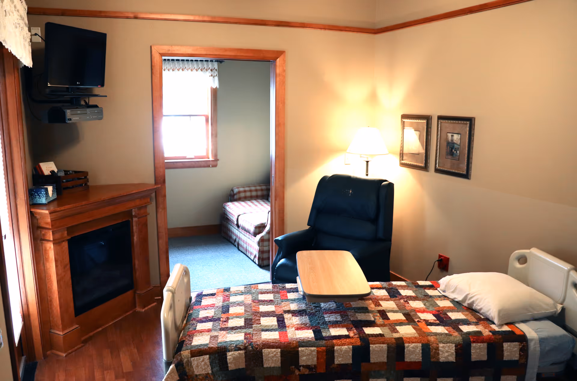 Patient bed with quilt, chair, small table, fireplace, and mounted TV in a cozy bedroom.