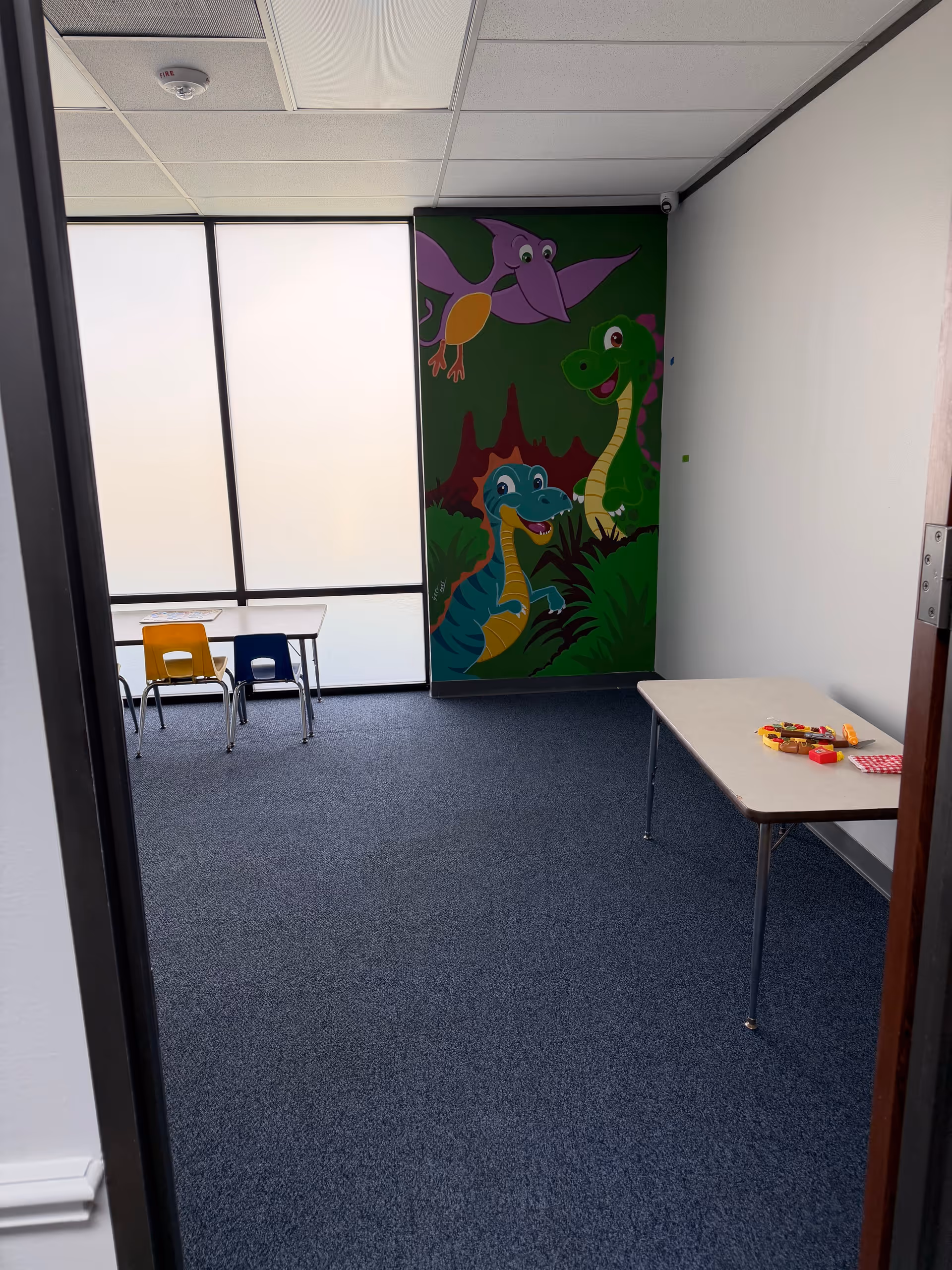 Interior activity room with a colorful dinosaur mural, small tables, and chairs.