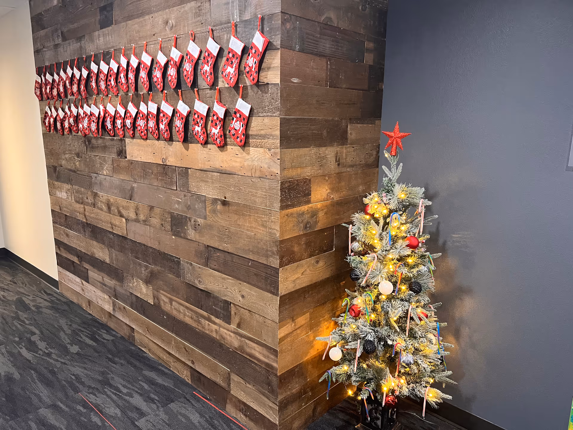 Corridor corner with rustic wood wall, rows of red stockings, and a lit Christmas tree.