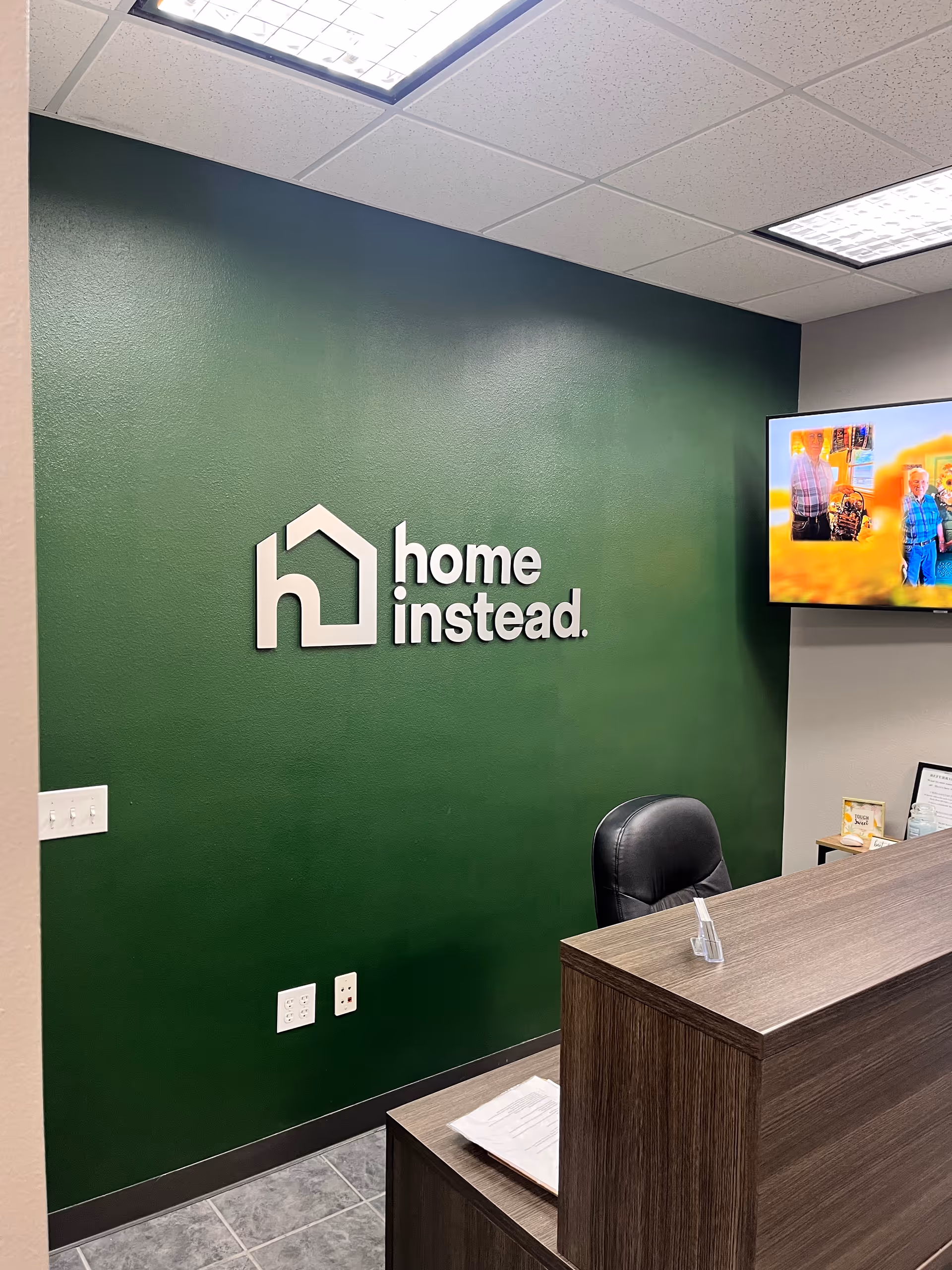 Reception desk in Home Instead lobby with green wall, logo, and TV.