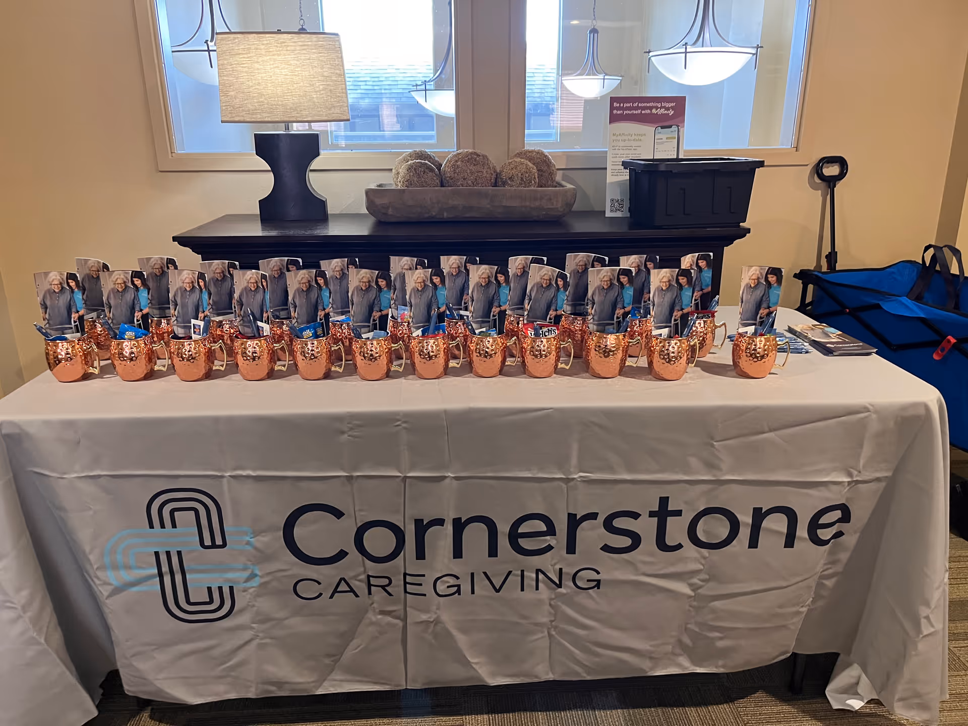 Indoor display table featuring Cornerstone Caregiving branding, copper mugs, and photo cards.