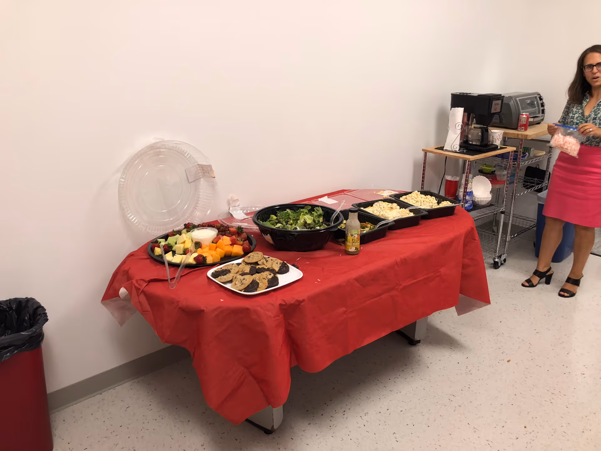 Buffet-style table with fruit, salad, and cookies; a staff member stands nearby.
