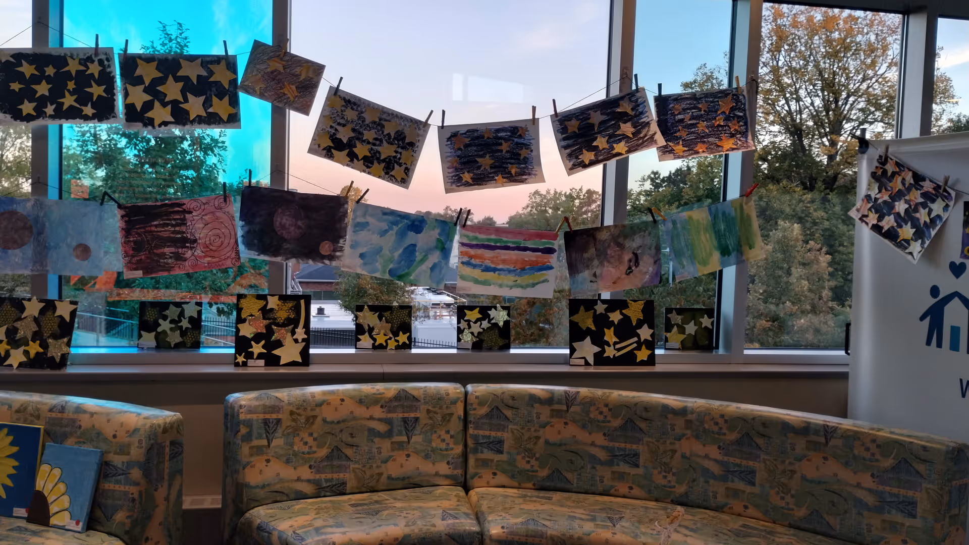 Colorful children's artwork hangs on a clothesline across a bright lounge with patterned sofas.