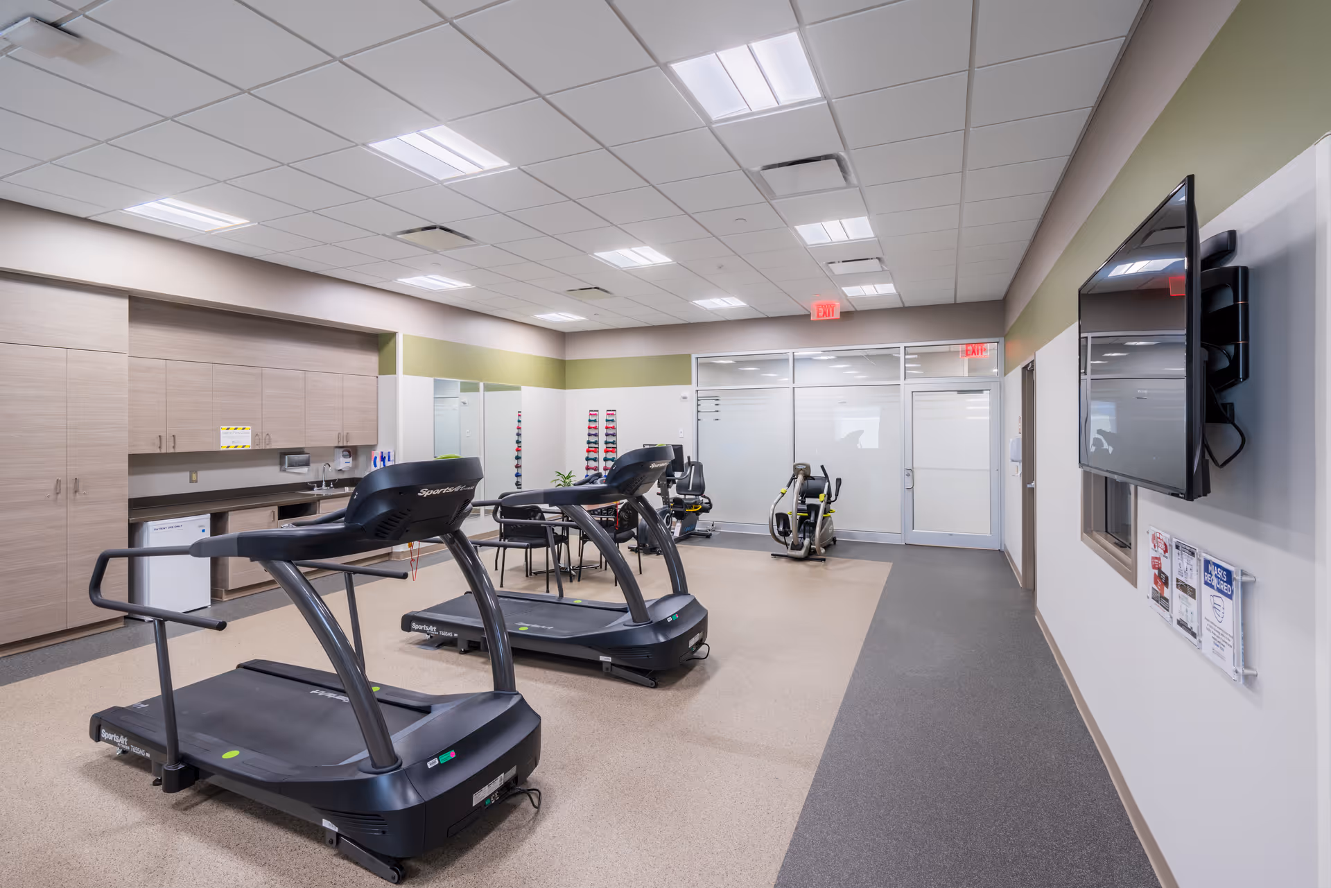 A modern rehabilitation gym with treadmills, exercise bikes, and a wall-mounted TV.