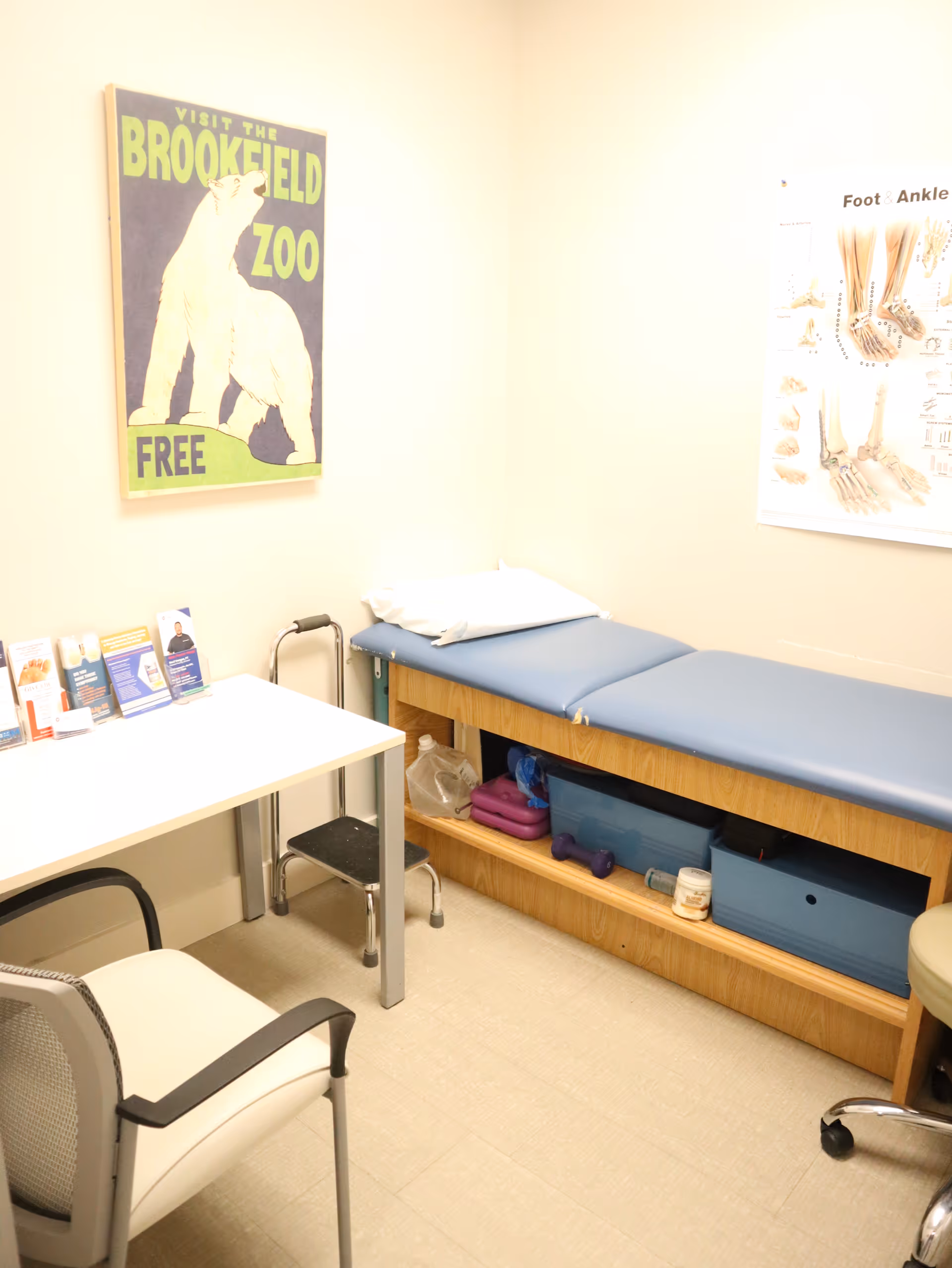 Clinic exam room with blue padded examination bed, desk, brochures, and posters.