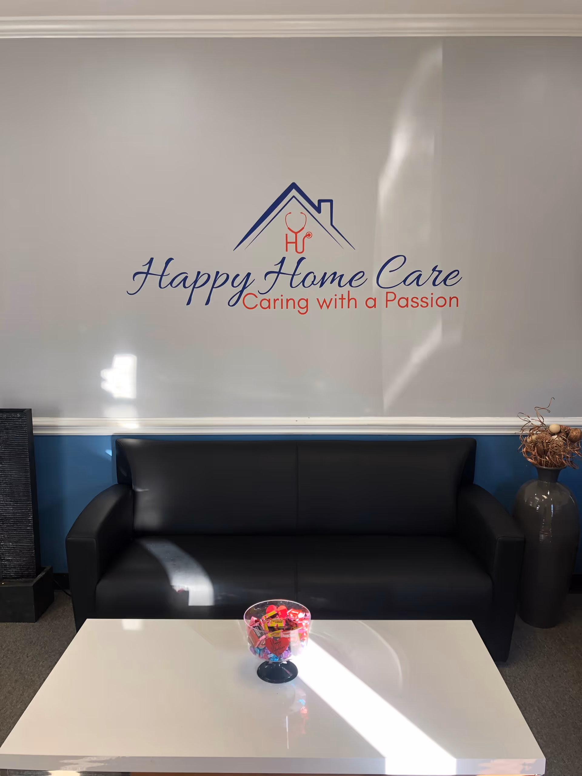 Lobby reception with a black sofa, white coffee table, and Happy Home Care logo on wall.