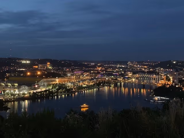 Nighttime cityscape along a river with illuminated buildings and reflections on the water.