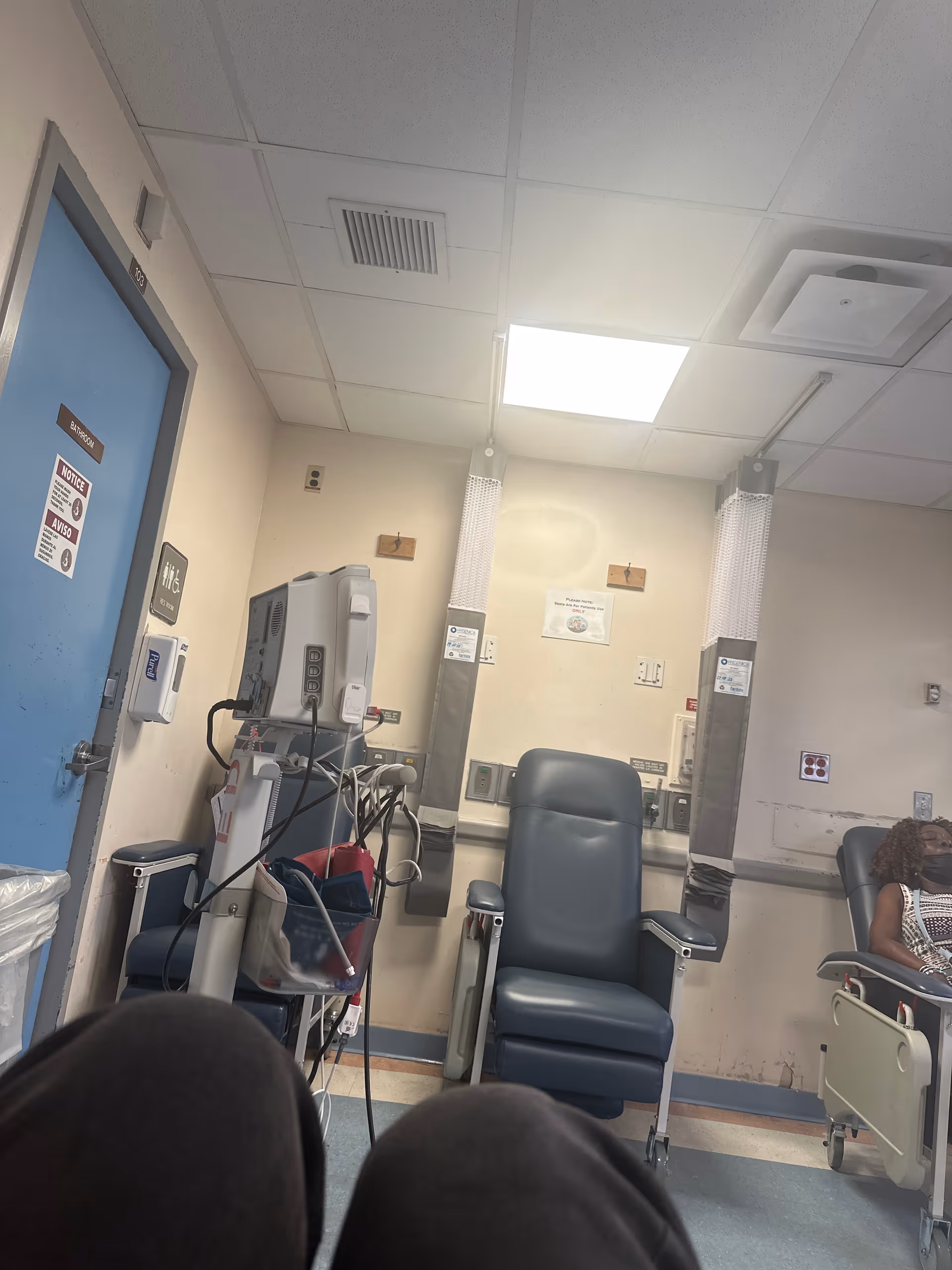Hospital treatment room with medical chairs, equipment, and a patient seated on the right.
