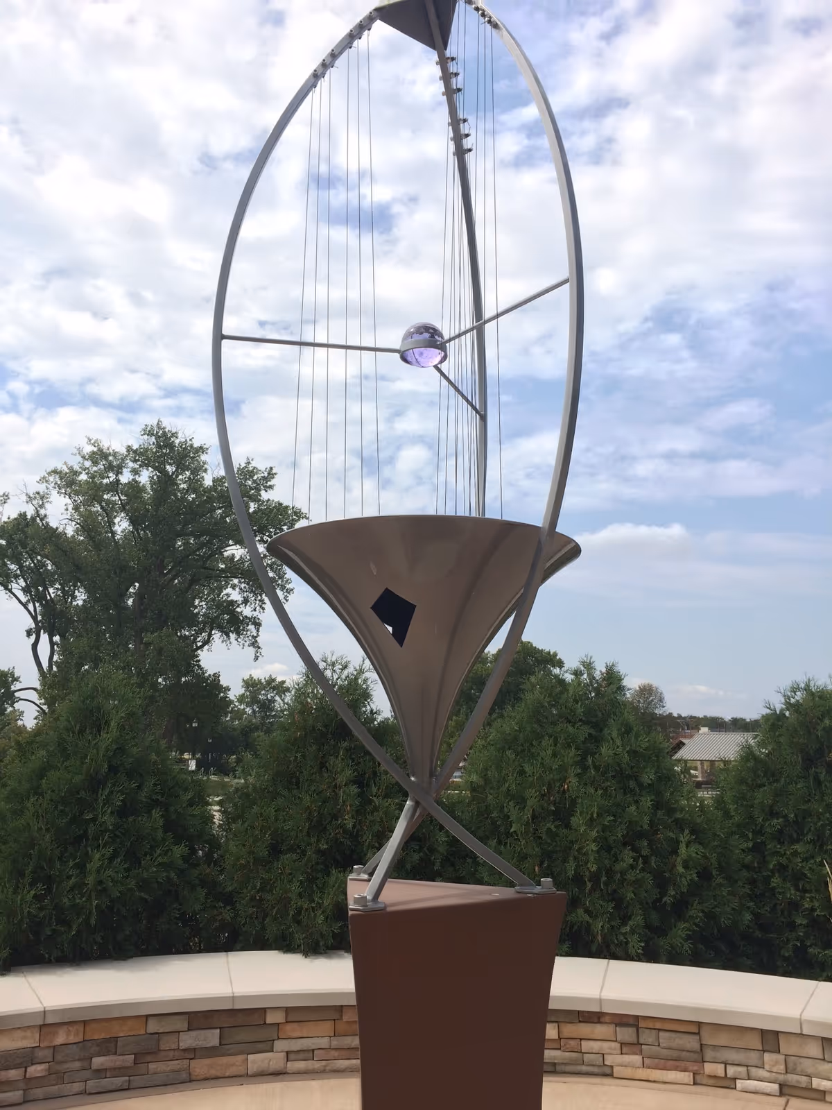Contemporary metal sculpture on a pedestal outdoors with trees and blue sky.