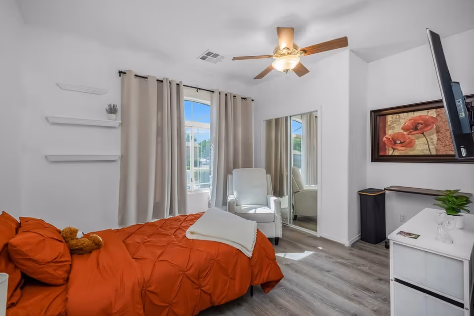 Bright bedroom with orange comforter, white furniture, and ceiling fan in Sunnyvale Group facility.
