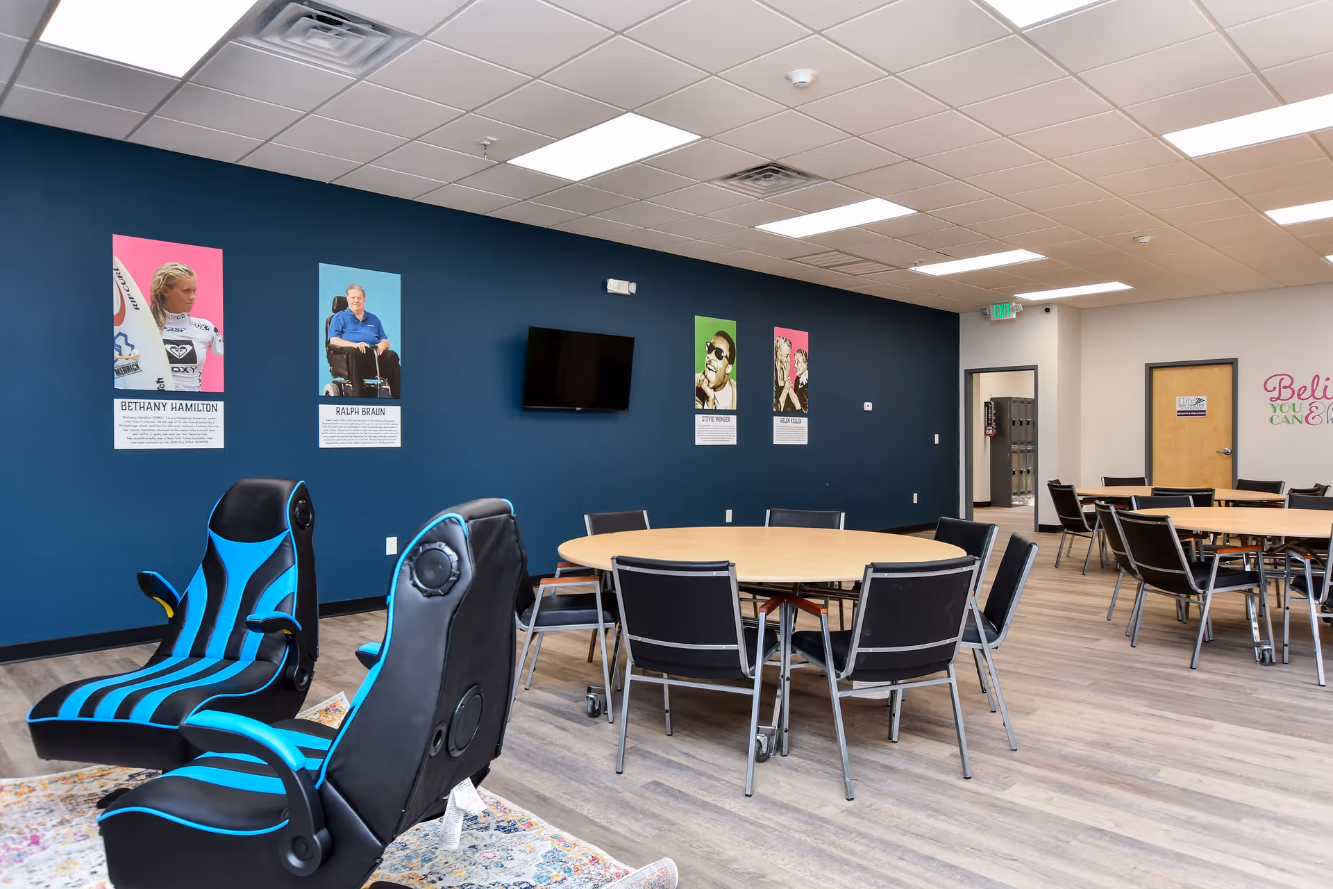 Interior of a senior living facility common area with round tables, chairs, and posters.
