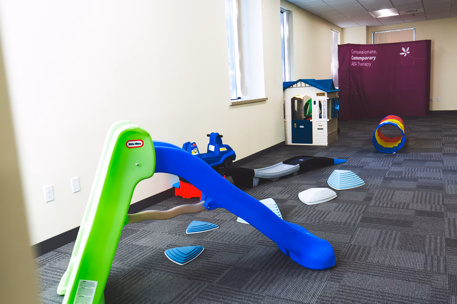 Bright indoor play area with colorful plastic slides, toy house, and tunnel in a senior facility.