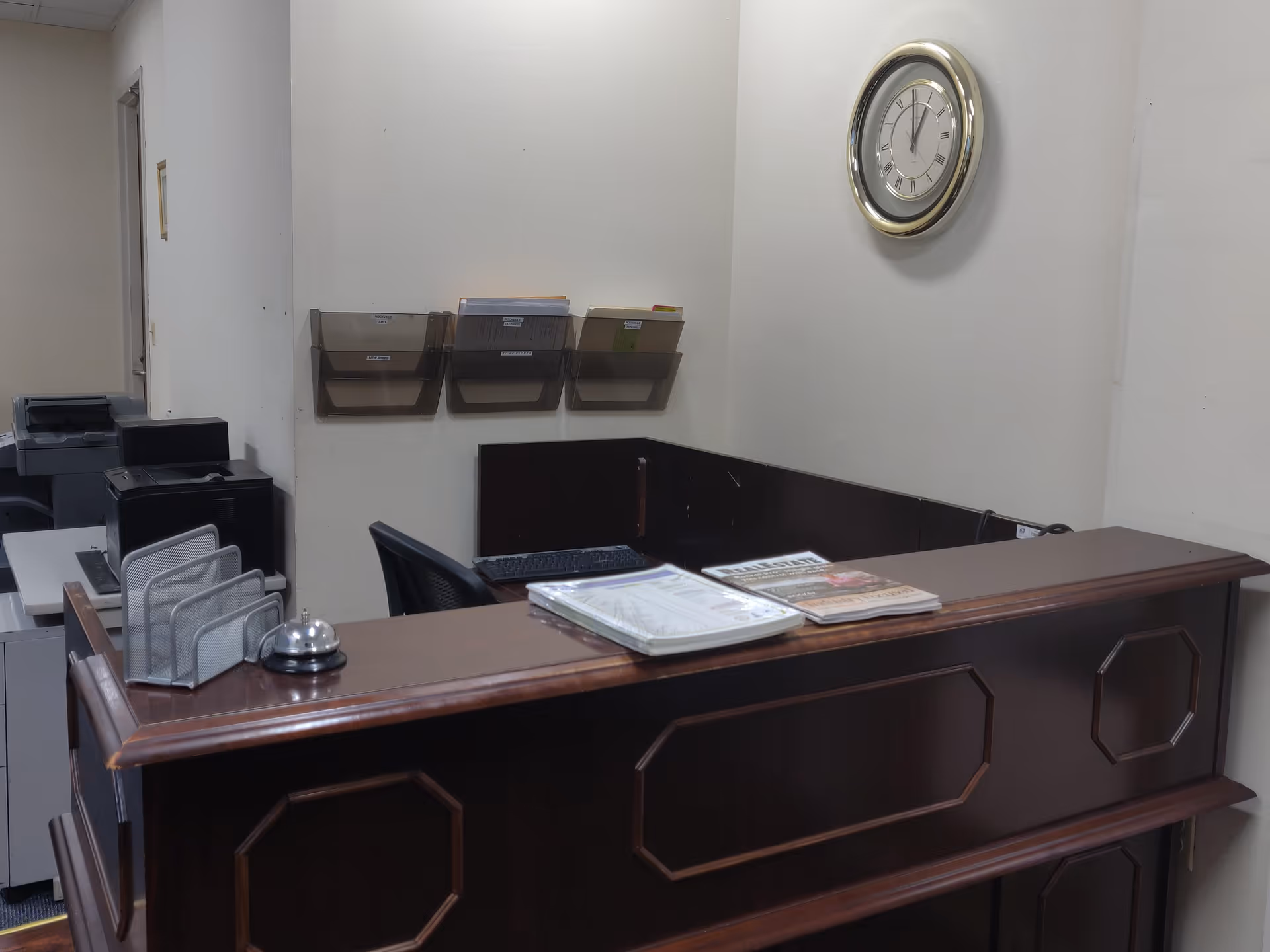 Dark wooden reception desk with magazines, mail slots, computer, and wall clock.