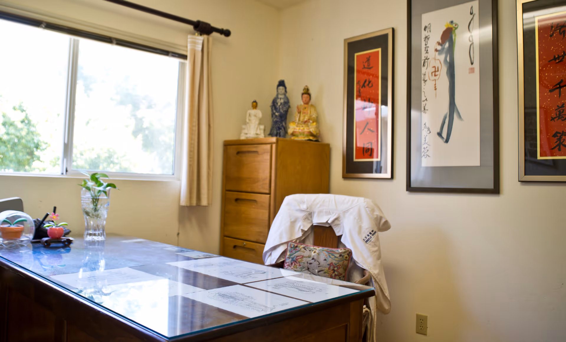 Office-style room with glass desk, plants, framed art, statues, and a chair draped with a coat.