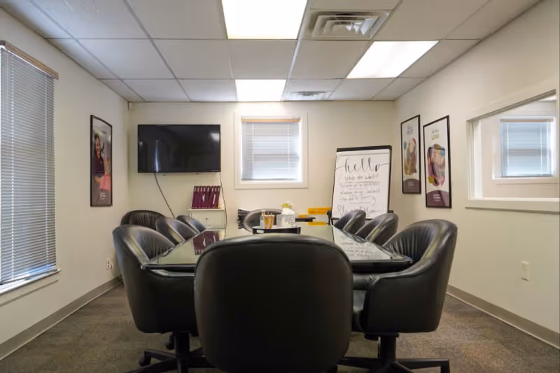 A conference-style meeting room with a long glass table, black chairs, and wall-mounted TV.