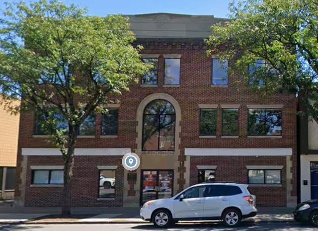 Brick two-story building facade with arched entrance, trees, and a white SUV parked outside.