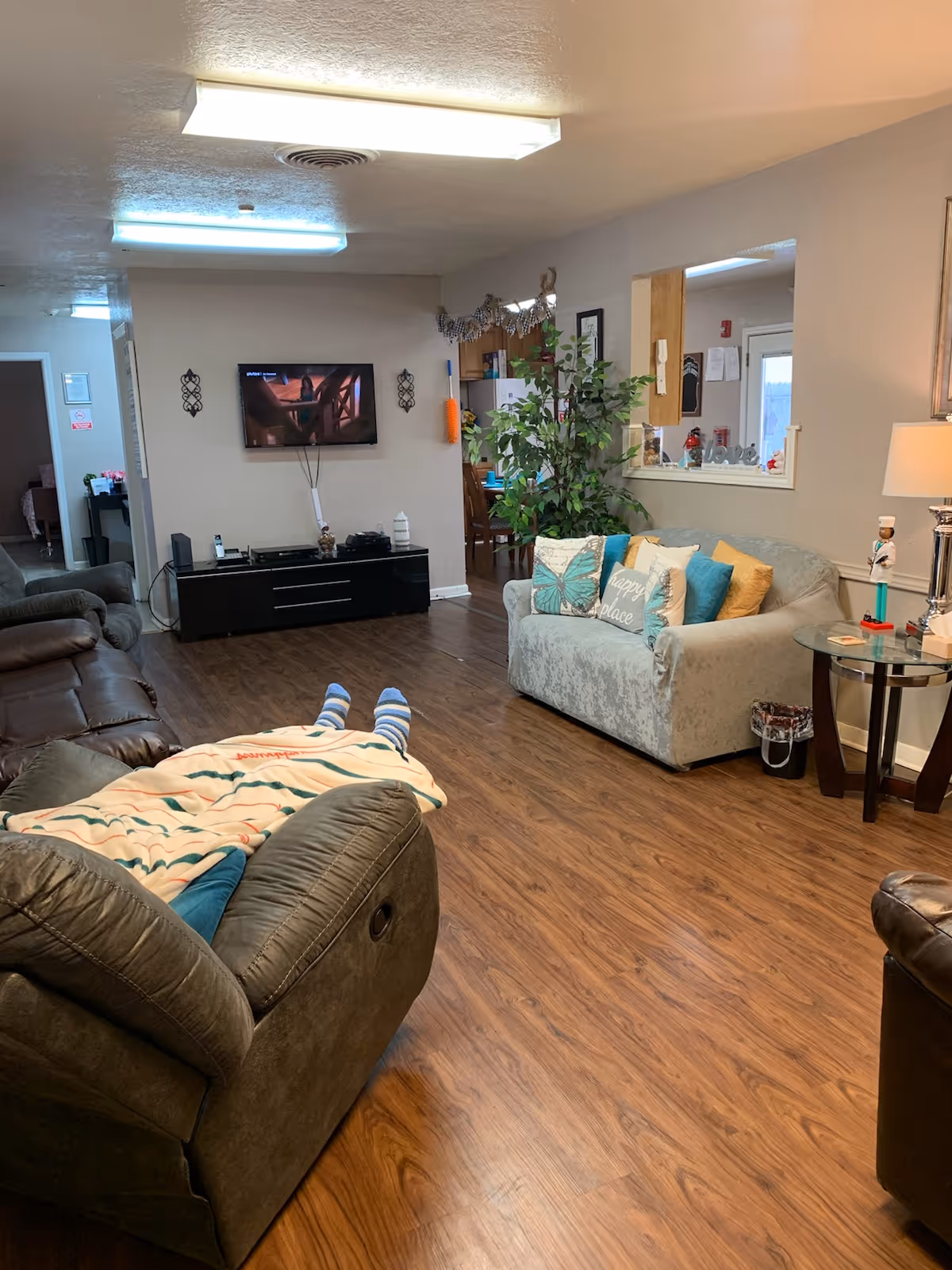 Cozy senior living room with sofas, TV, plants, and a resident resting under a blanket.