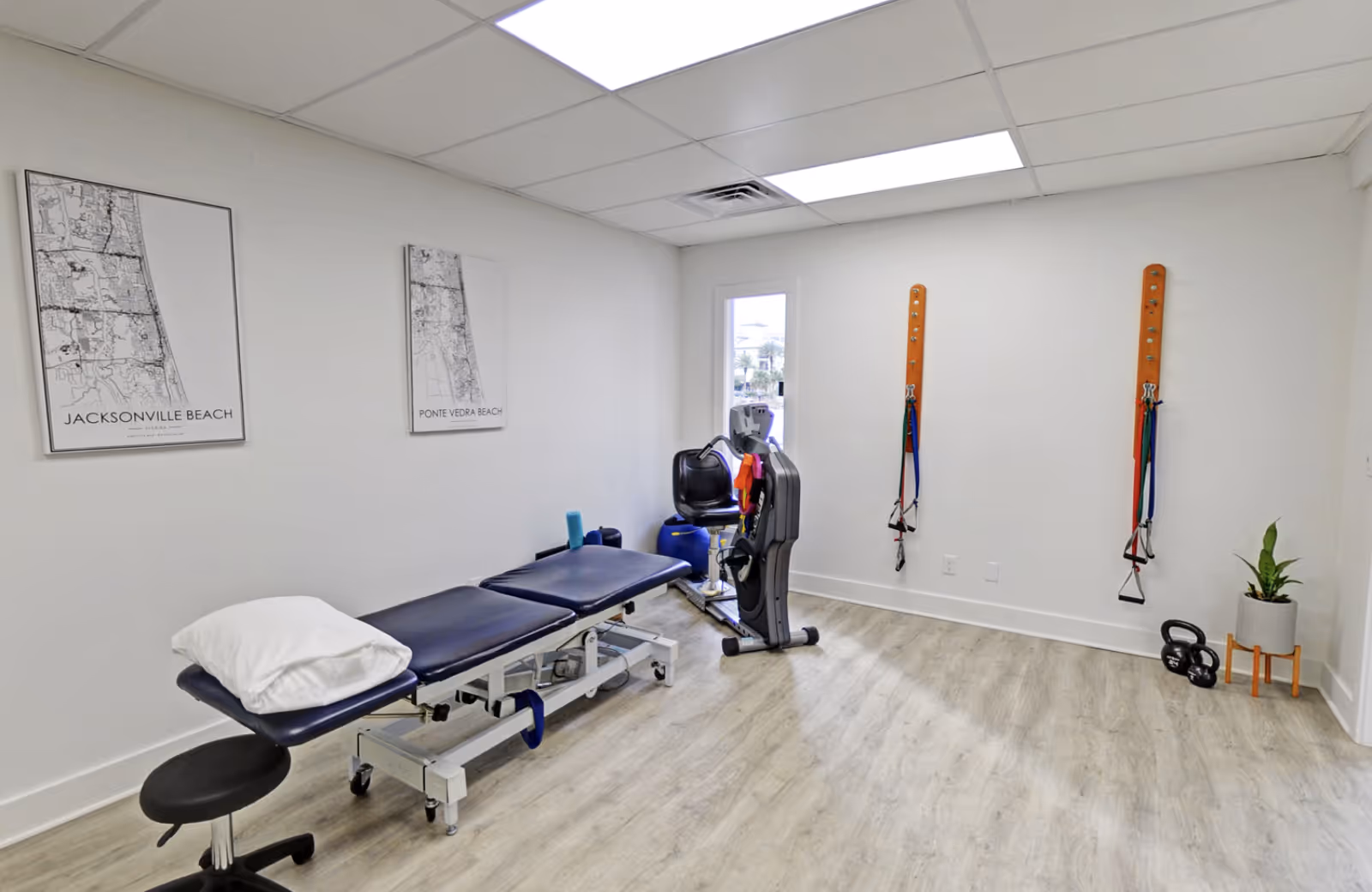 Therapy room with adjustable treatment table, exercise machine, wall maps, and resistance bands.