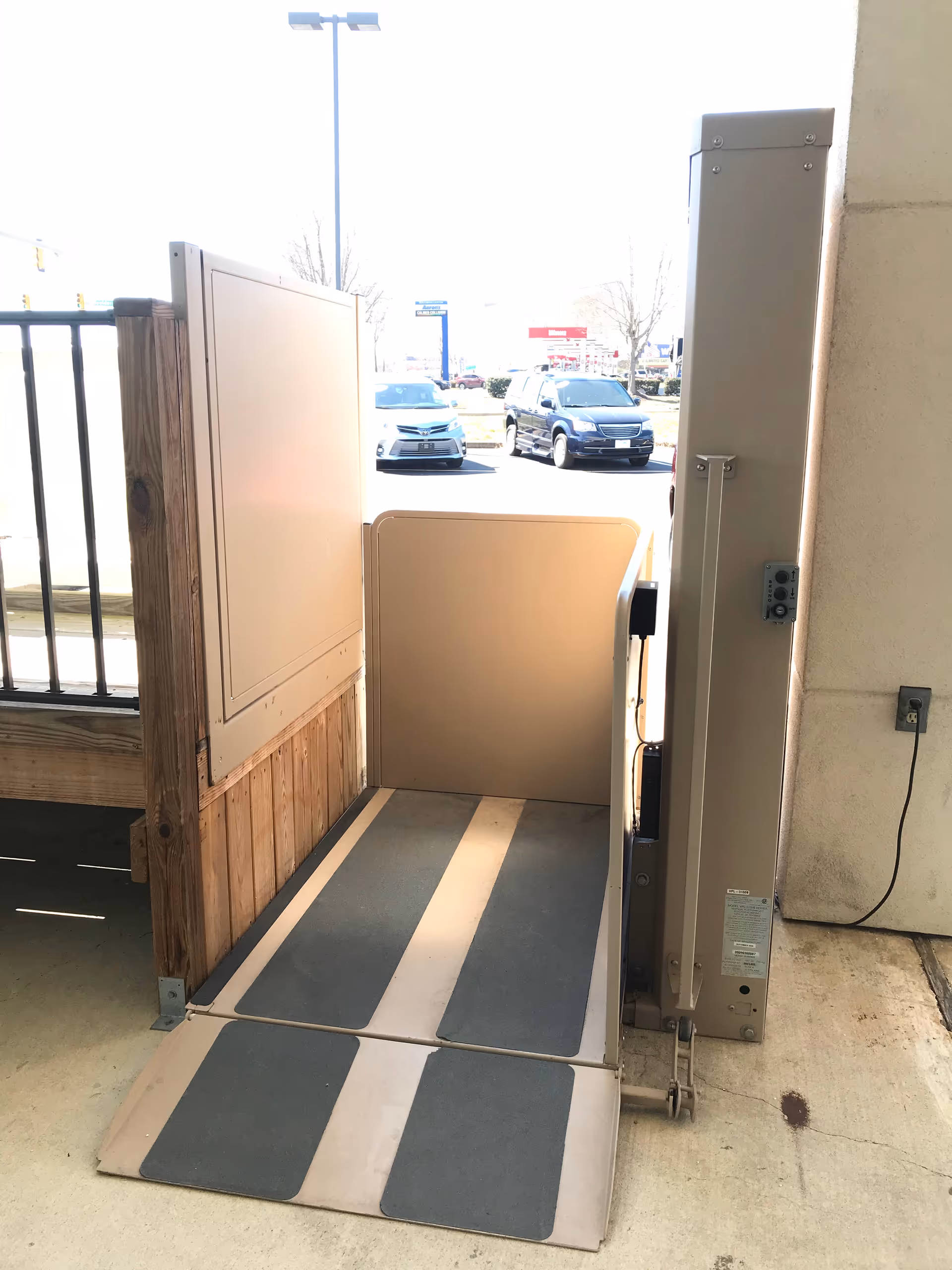 Exterior wheelchair accessibility lift at building entrance with ramp; parking lot visible beyond.