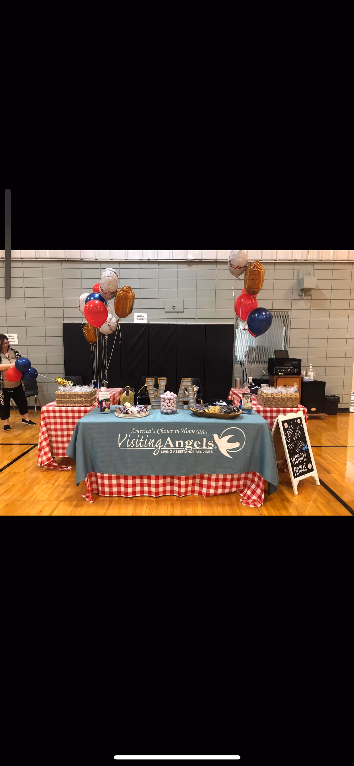 A fundraising/event booth with balloons and a Visiting Angels banner in a gym hall.