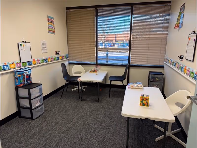 Small therapy room with two white tables, chairs, blinds, and an educational wall border.