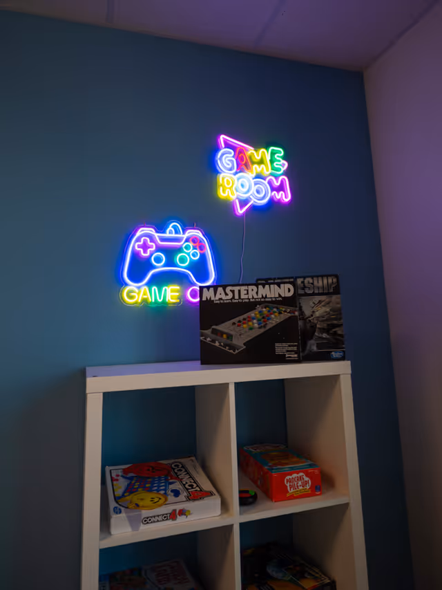 Blue wall with a neon GAME ROOM sign above a shelf of board games.