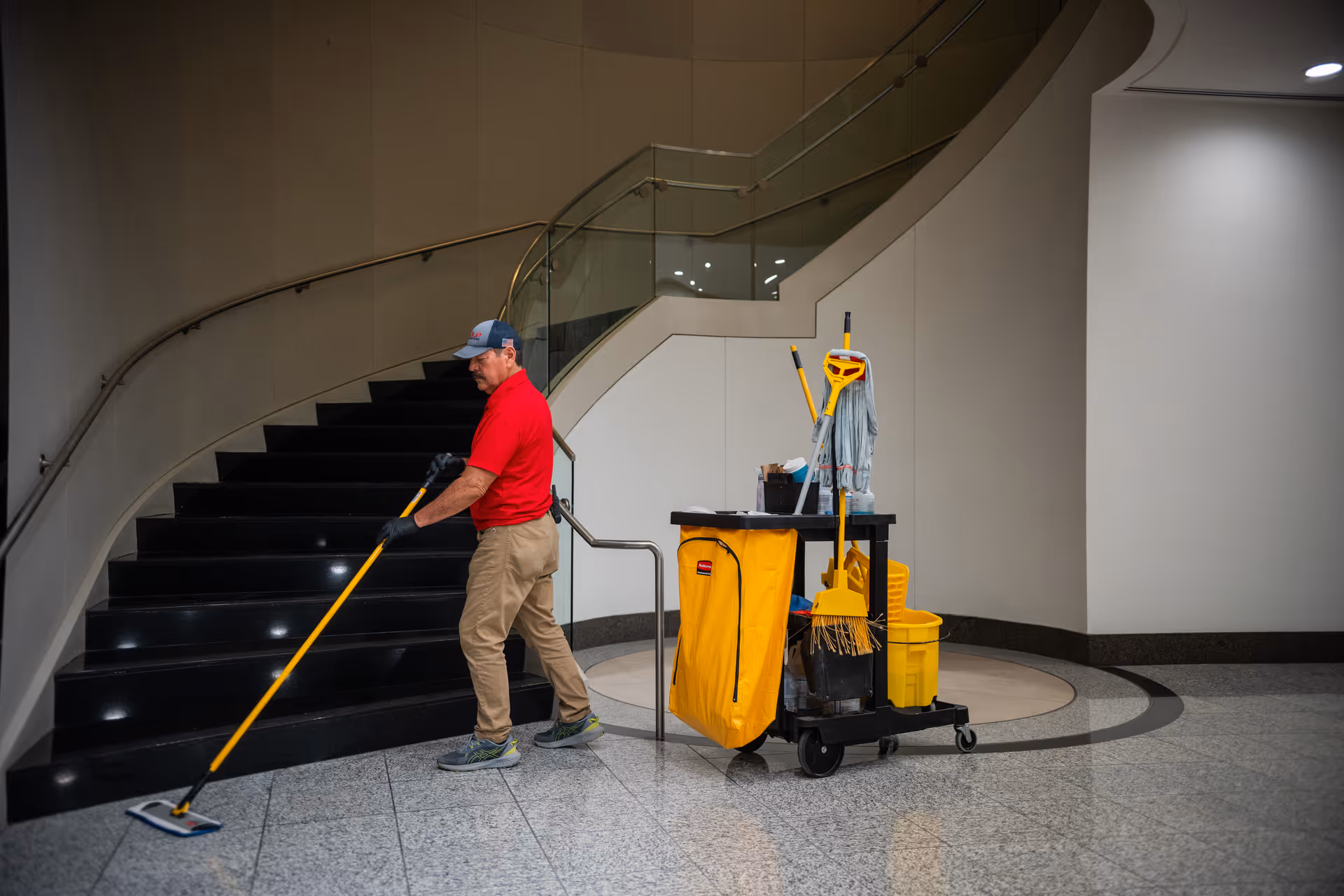 Maintenance worker in a red shirt mops the lobby floor beside a cleaning cart.