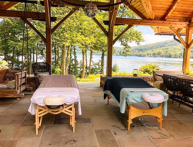 Outdoor covered patio with two massage tables overlooking a lake and trees.