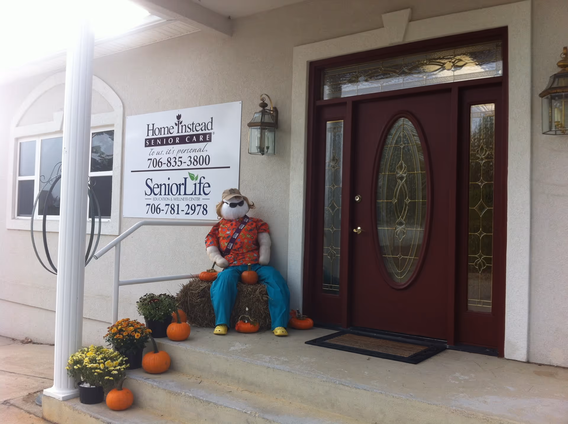 Front entrance of Home Instead Senior Care with a scarecrow statue, pumpkins, and potted plants.