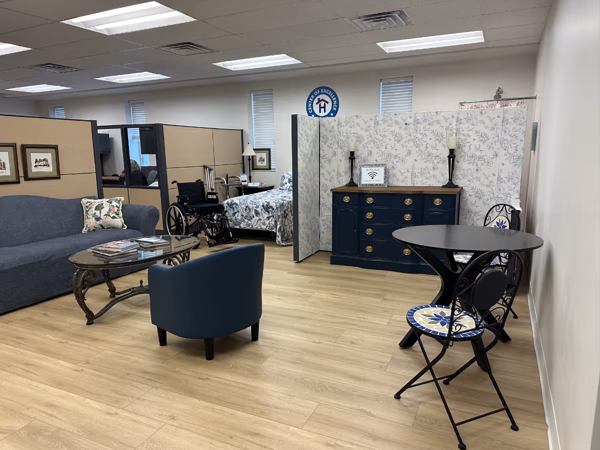 Bright senior living lounge with sofa, chairs, coffee table, and a bedroom area visible in back.