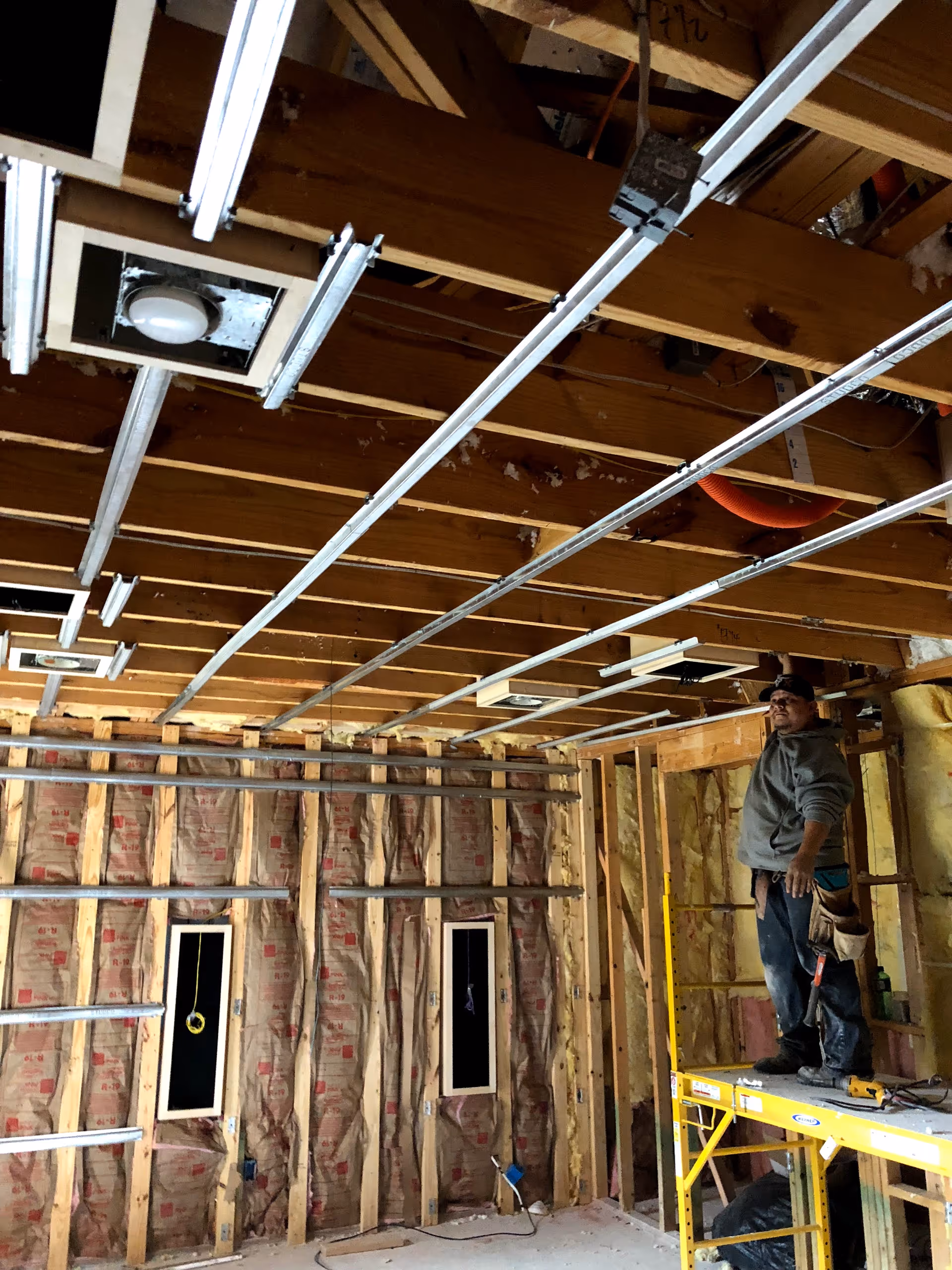 Interior construction site with exposed framing, pipes, and a worker on scaffolding.