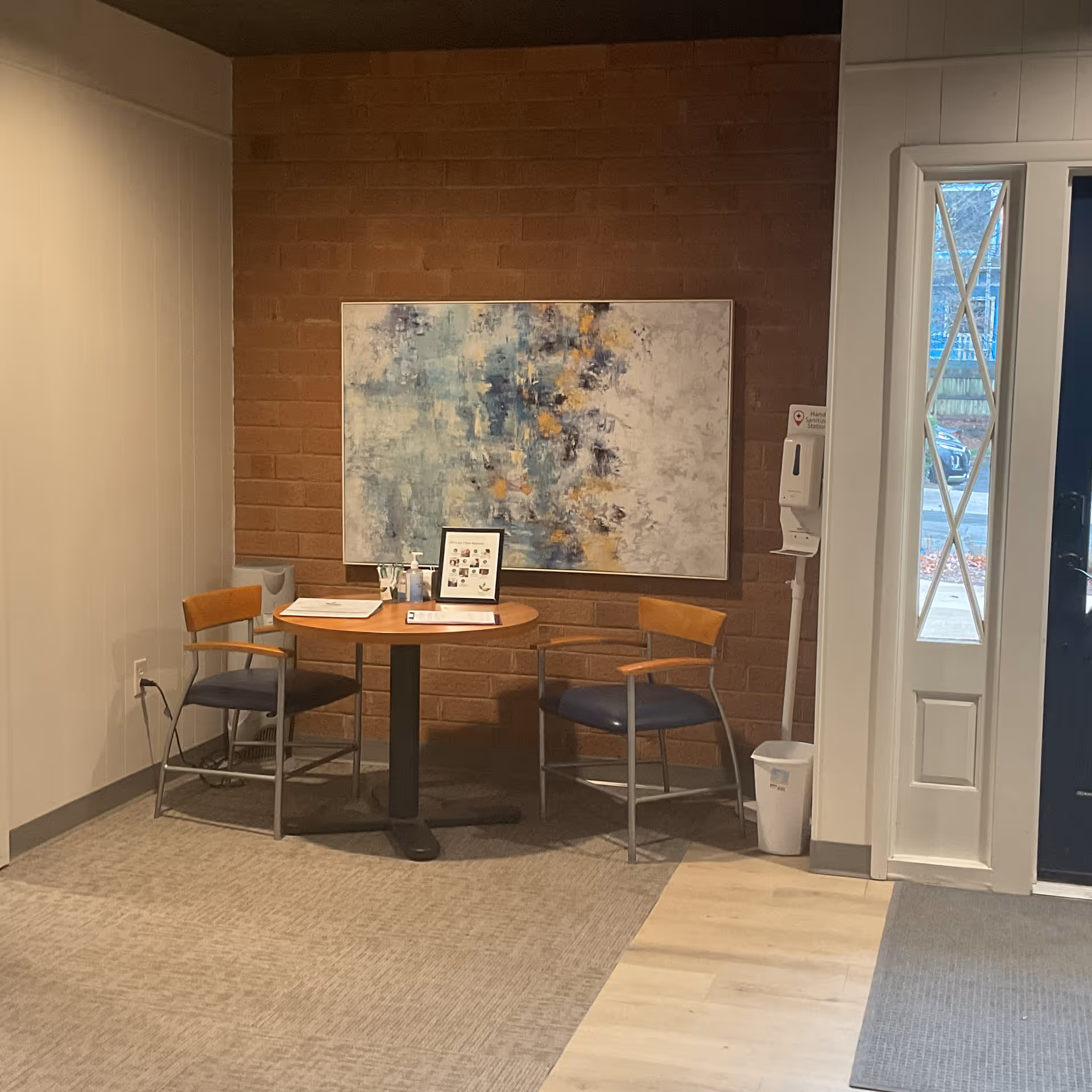 A small seating area with a round table, two chairs, artwork, and a hand sanitizer station.