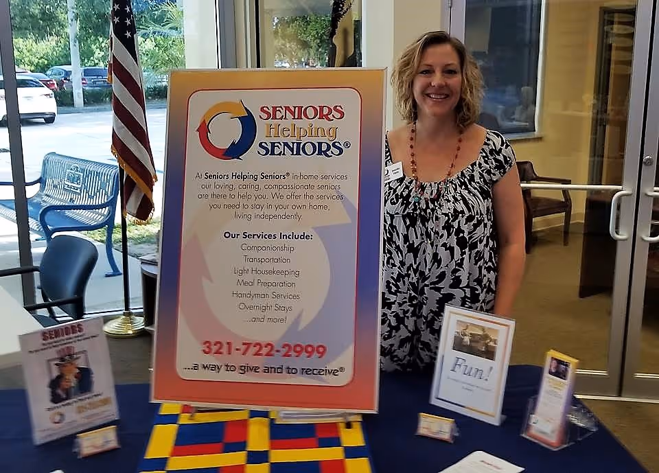 Smiling woman stands beside a large Seniors Helping Seniors banner in a facility lobby.