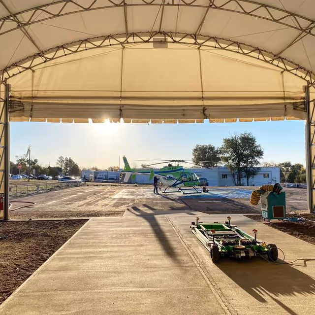 A green helicopter sits on a tarmac outside a large white tent shelter.