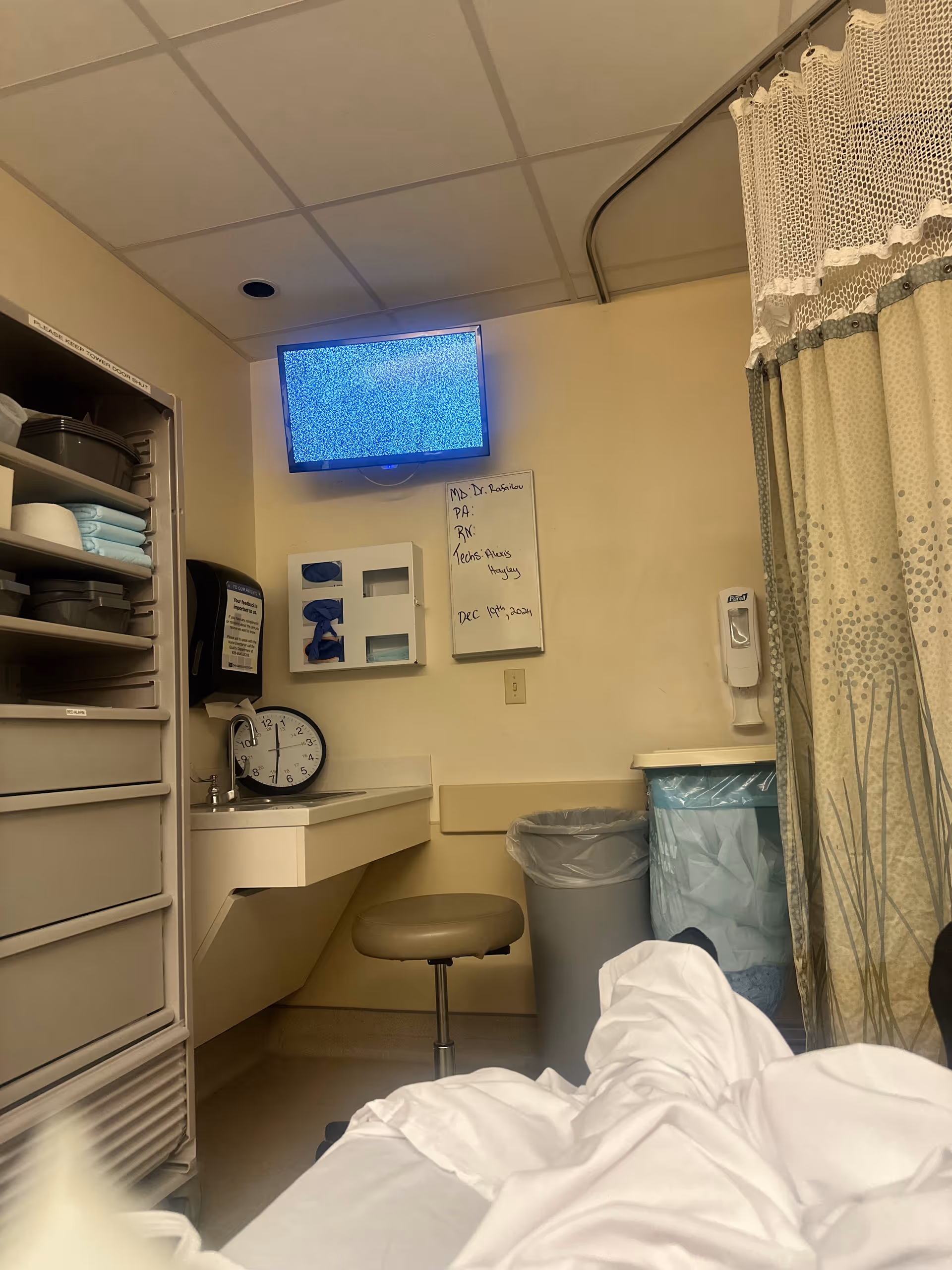 A hospital-style patient room with a blue static monitor, sink, shelves, and curtain.