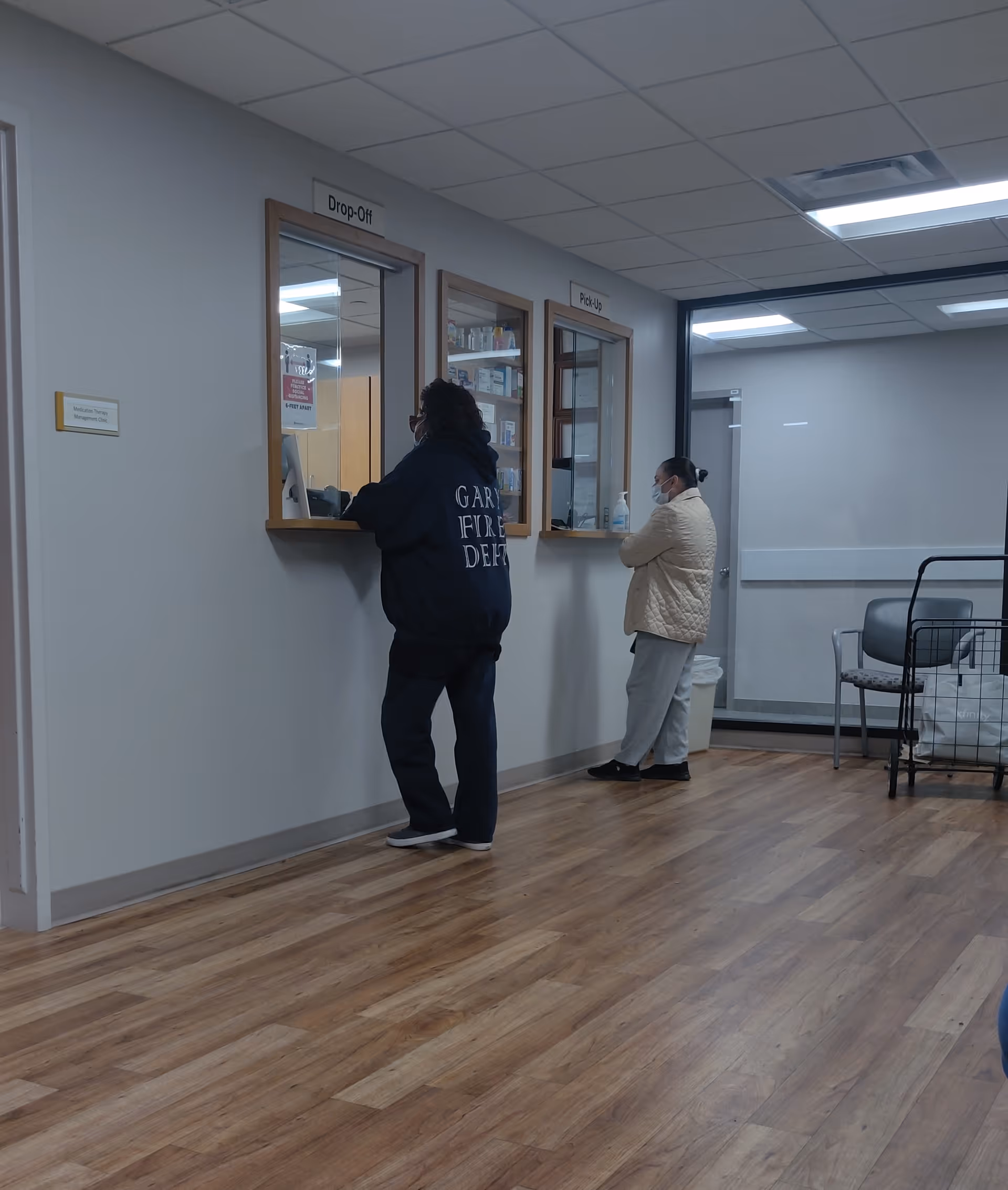 Two people stand at service windows in a hospital-like interior corridor.