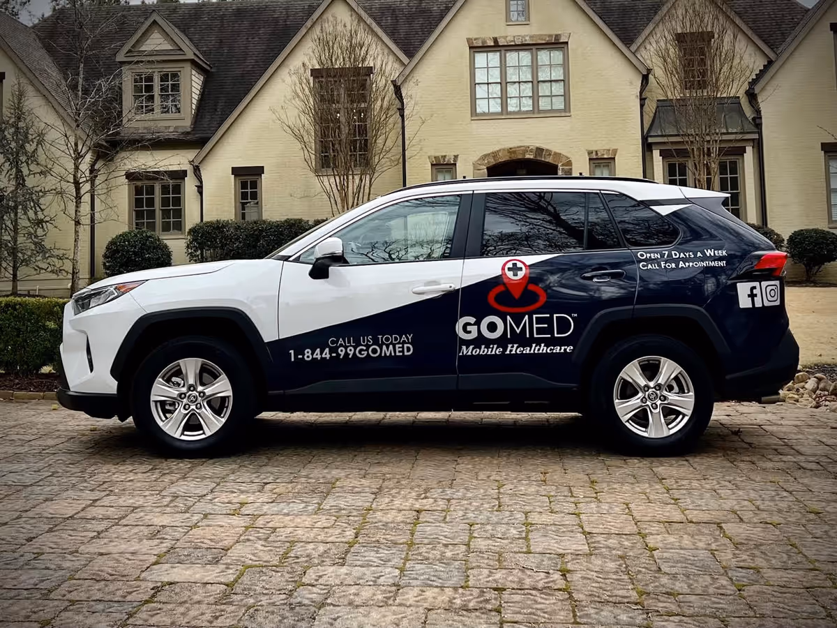 White and dark blue GOMED Mobile Healthcare SUV parked in front of a beige brick house.