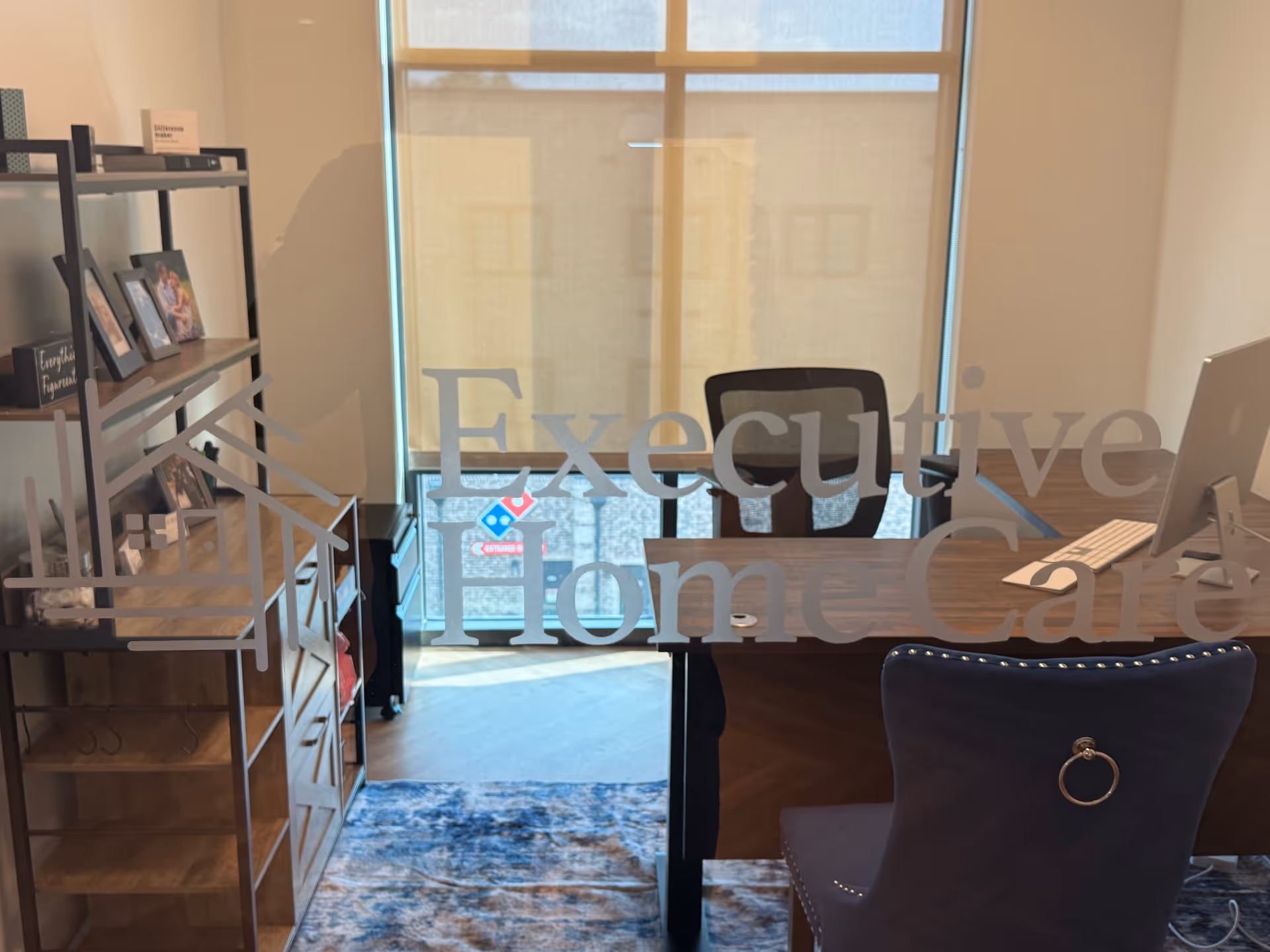 Office reception area with glass logo, desk, chair, and shelving inside a senior care facility.