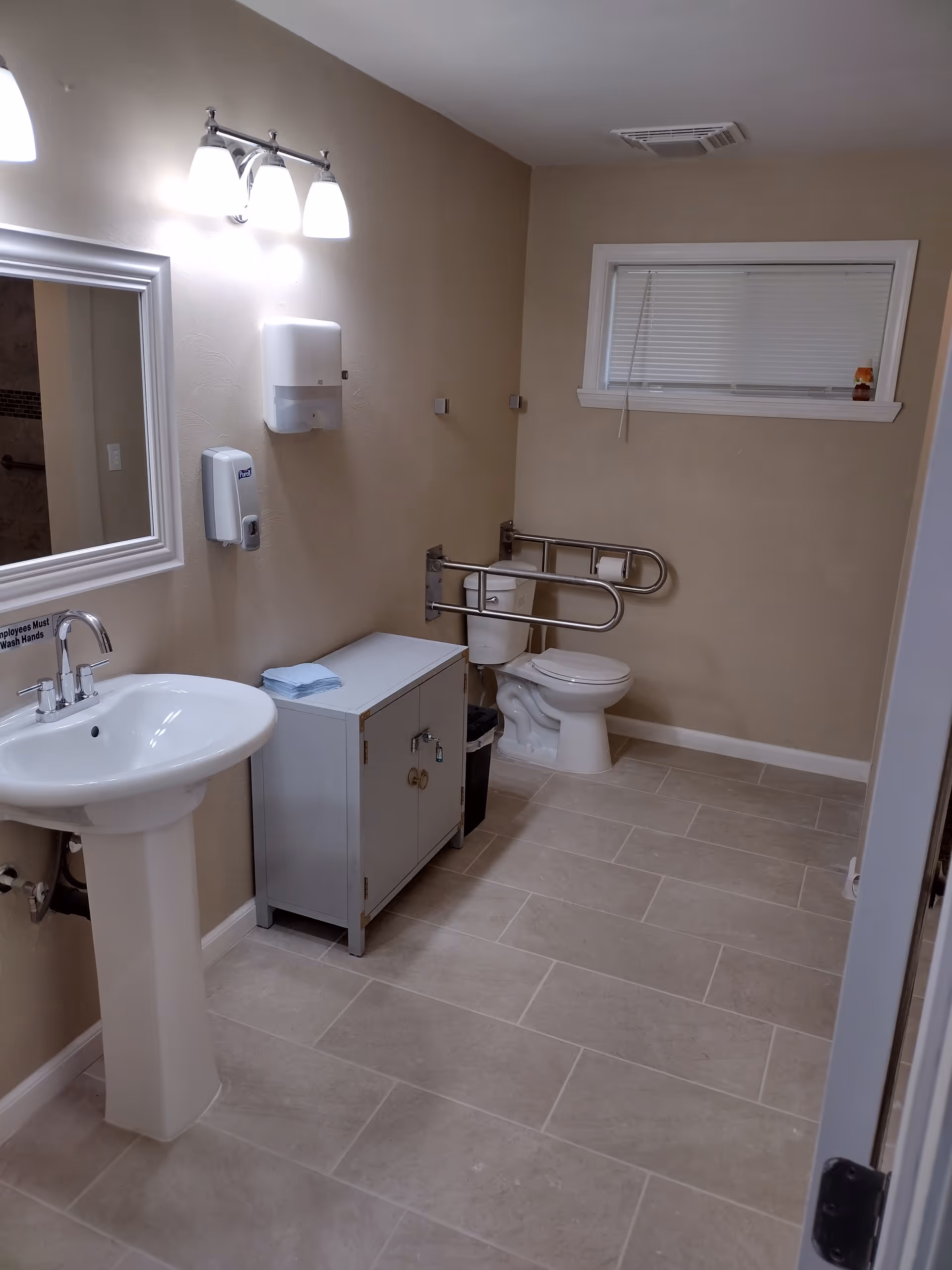 Accessible bathroom with pedestal sink, cabinet, toilet with grab bars, wall mirror, and window.