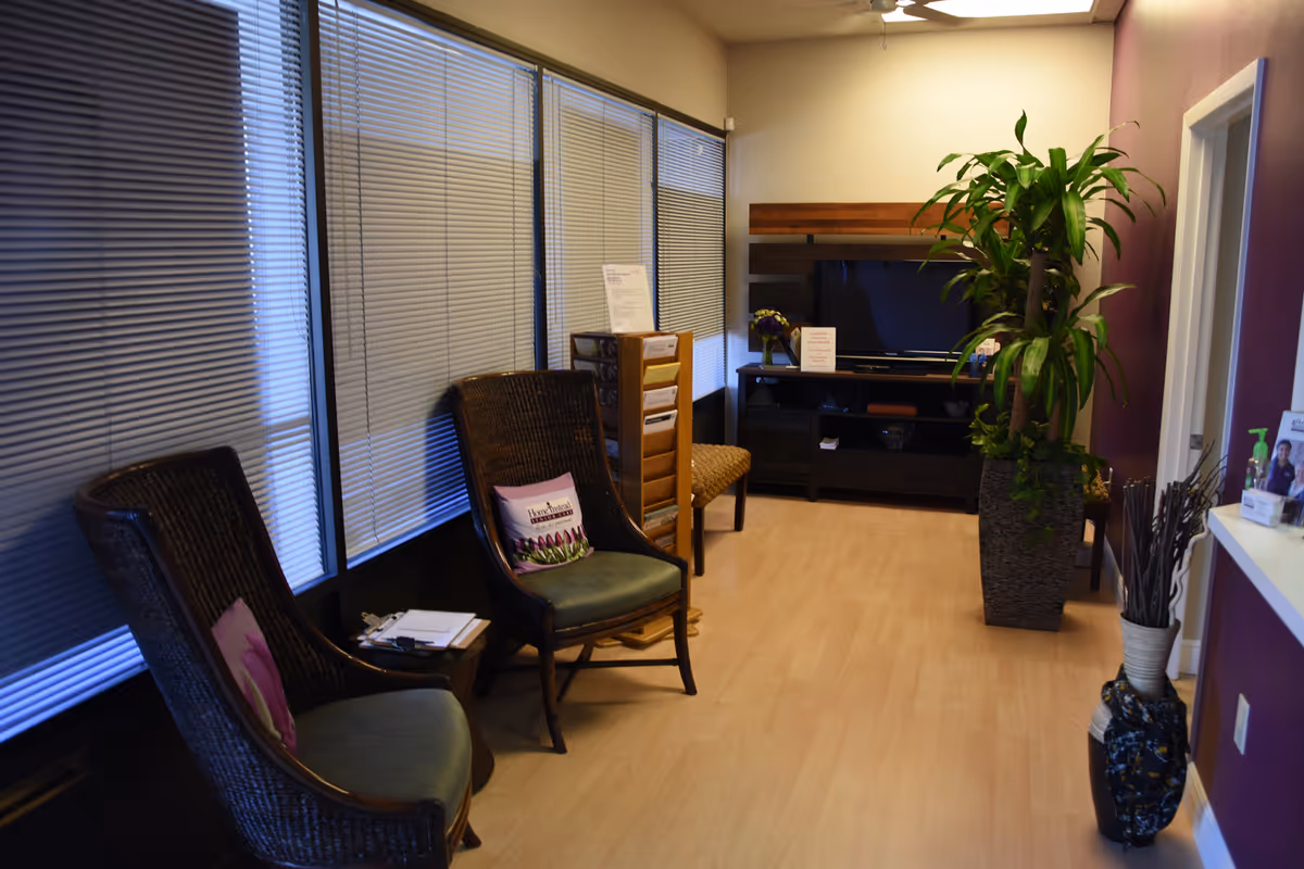 An interior waiting area with wicker chairs, blinds, a TV stand, and a tall plant.