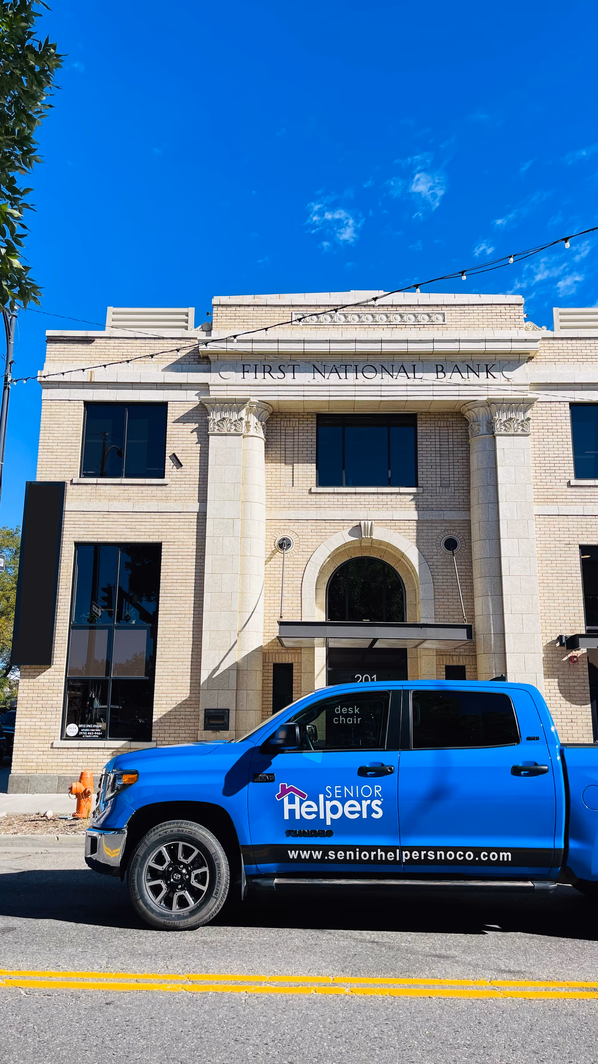 Blue Senior Helpers van parked in front of a beige bank building on a sunny day.
