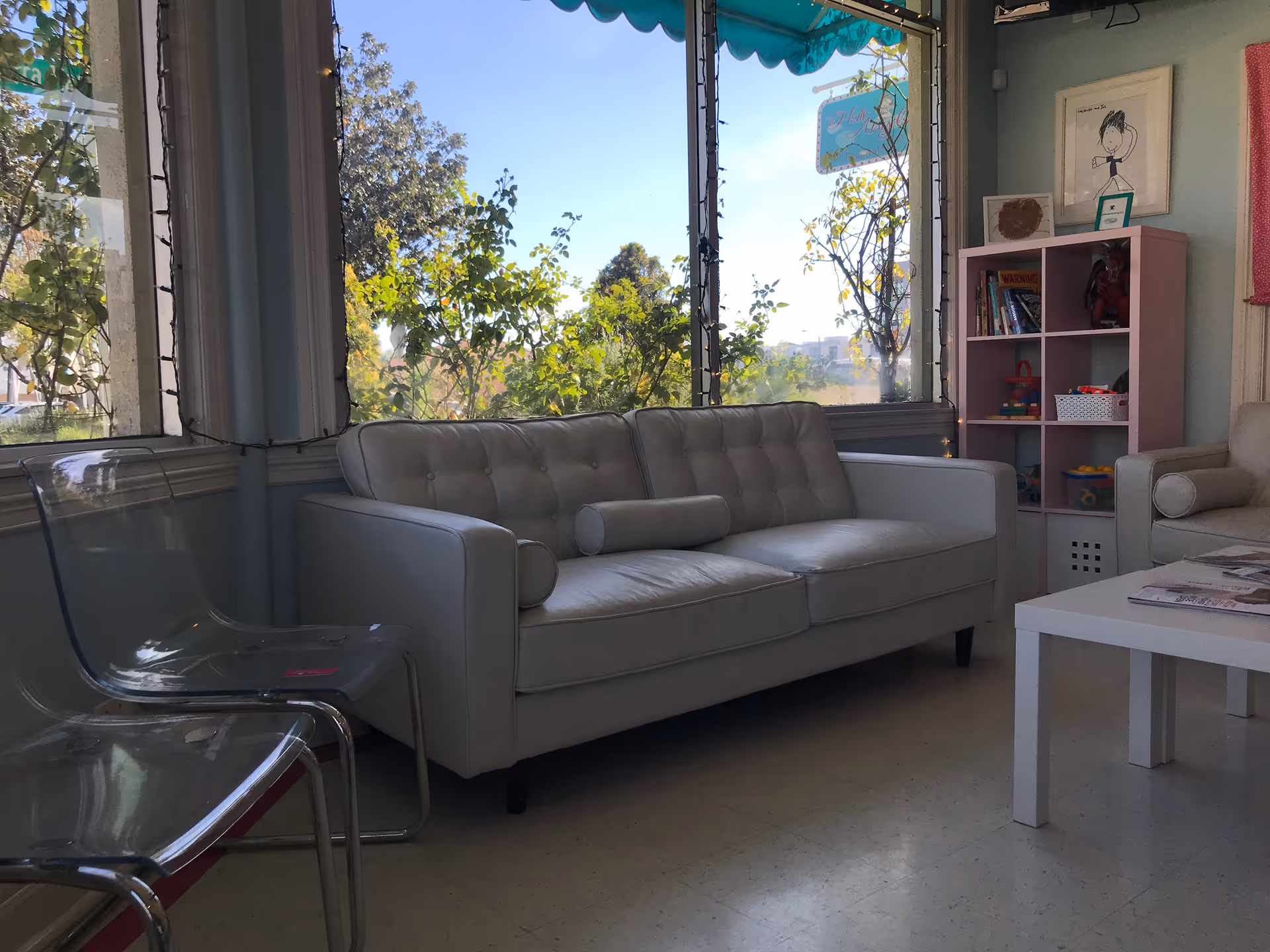 Bright indoor lounge with a light-gray sofa, white coffee table, and toy shelf near windows.