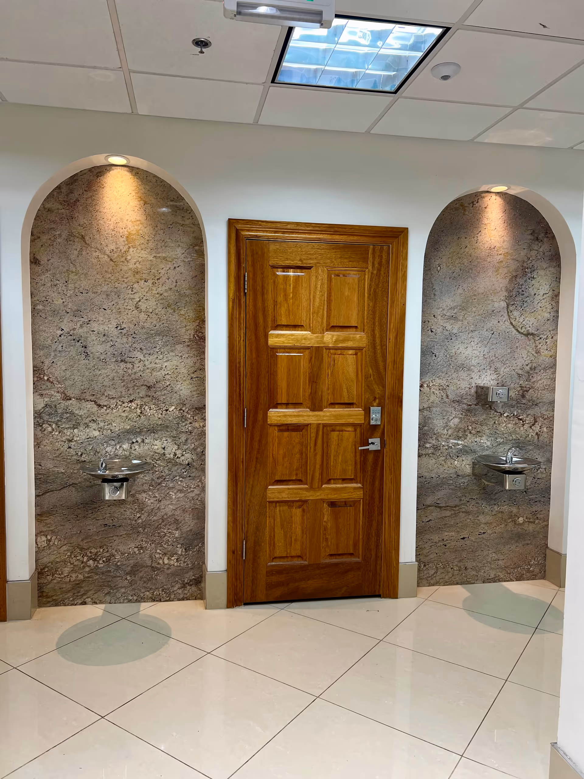 Wooden door between two marble arches featuring wall-mounted sinks in a bright interior hall.
